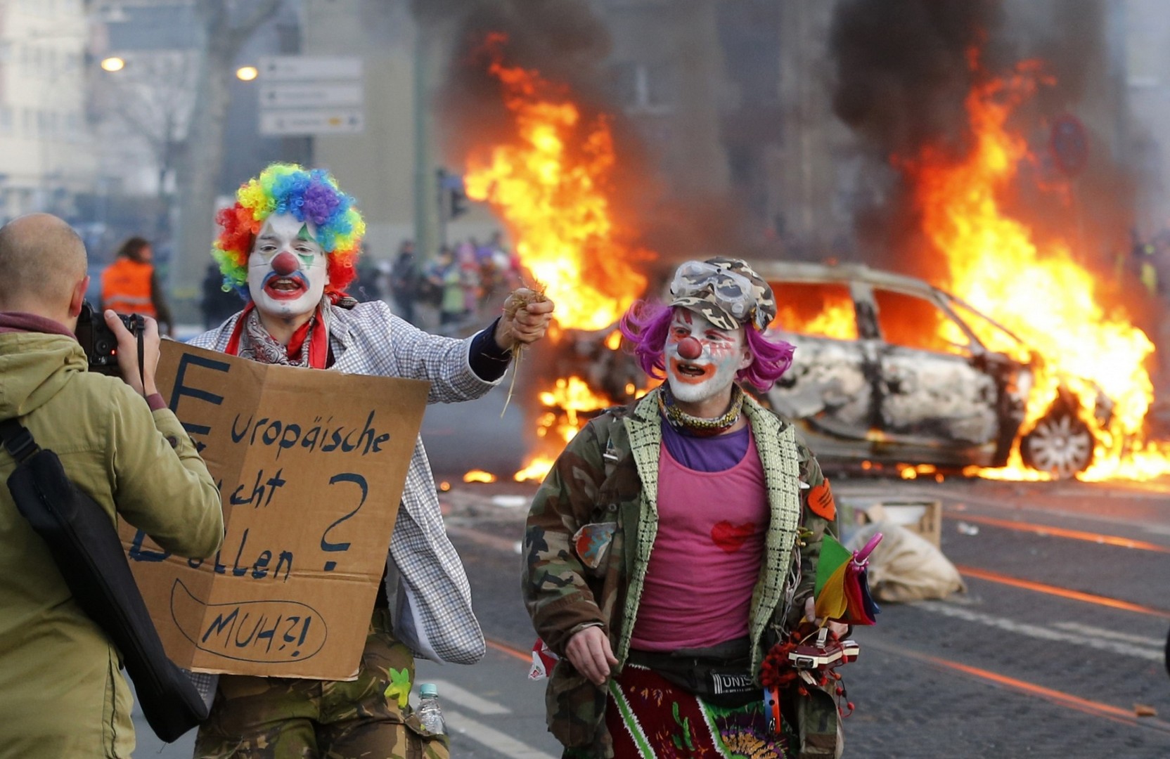 Διαδηλωτές στη Φρανκφούρτη ντυμένοι κλόουν με φόντο σοβαρά επεισόδια στο κέντρο της πόλης.